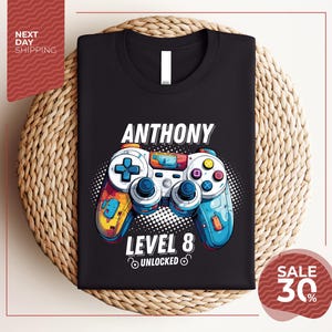 Könnte beinhalten: Schwarzes T-Shirt mit einem farbenfrohen Videospiel-Controller-Design. Der Aufdruck zeigt den Namen "Anthony" und den Text "Level 8 Unlocked". Der Controller hat blaue, gelbe und orangefarbene Akzente.