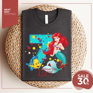 A gray t-shirt with a design featuring the character Ariel from the Disney movie "The Little Mermaid". The design includes the number 7 and the text "Birthday Girl Lucy".