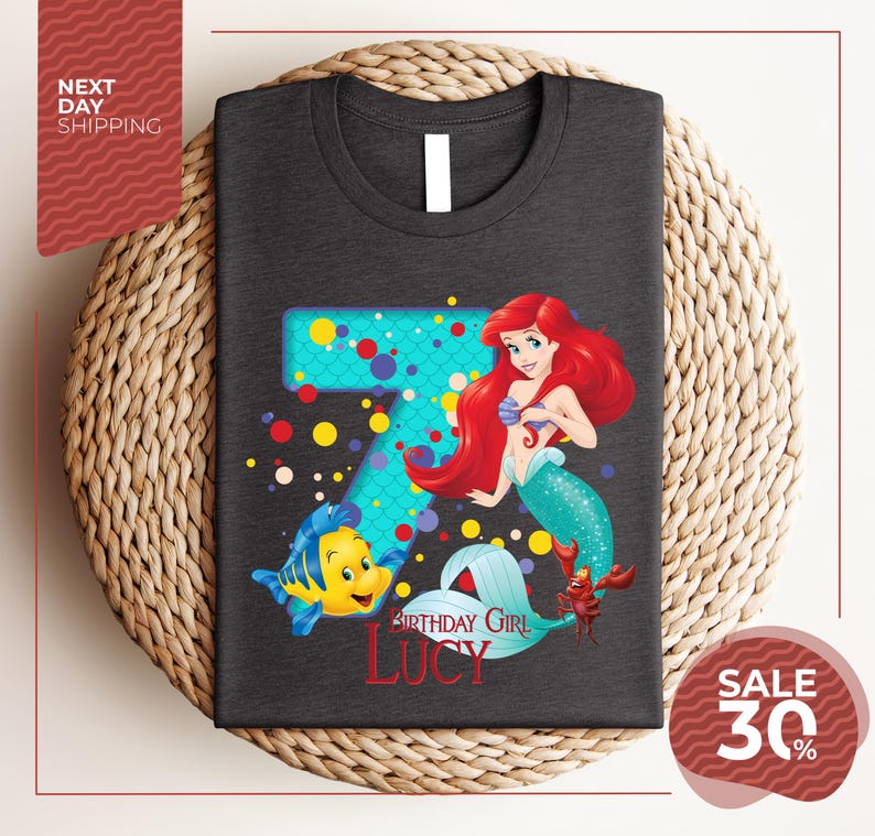 A gray t-shirt with a design featuring the character Ariel from the Disney movie "The Little Mermaid". The design includes the number 7 and the text "Birthday Girl Lucy".