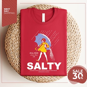 a red t-shirt with a graphic design depicting a woman holding a salt shaker and sprinkling salt on the ground, with the text "ALL these flavors and to be chosen" and "SALTY" below the image.