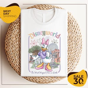 A white t-shirt with a Disney World-themed graphic design, featuring a cartoon image of a duck and various Disney landmarks.