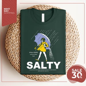 a green t-shirt with a graphic design depicting a person holding an umbrella and sprinkling salt on the ground. The text on the t-shirt reads "ALL these flavors and to be chosen to be SALTY".