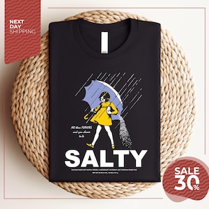 a black t-shirt with a graphic design depicting a person holding an umbrella and sprinkling salt on the ground. The text on the t-shirt reads "ALL these flavors and to be chosen to be SALTY".