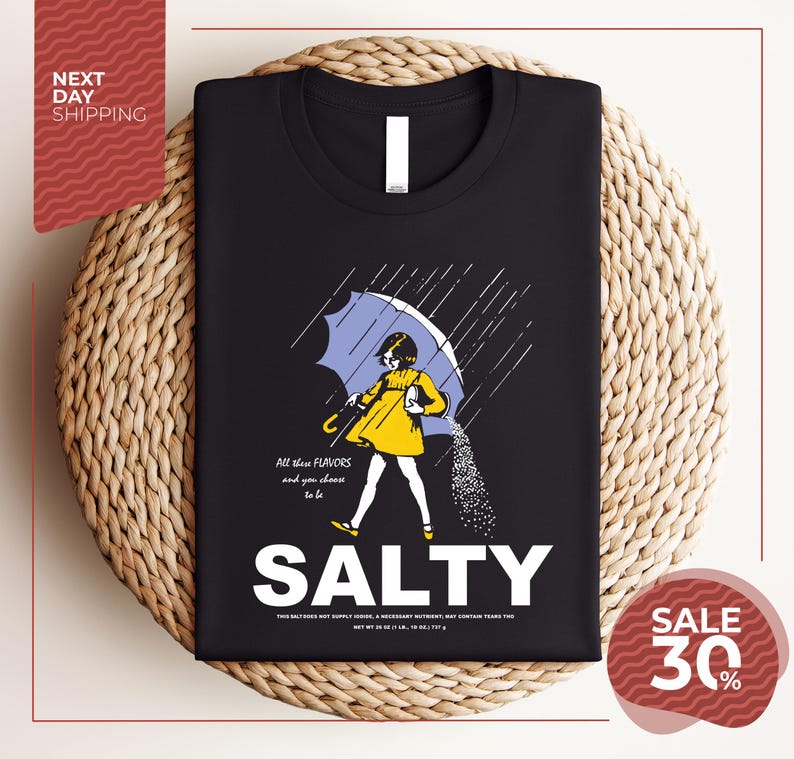 a black t-shirt with a graphic design depicting a person holding an umbrella and sprinkling salt on the ground. The text on the t-shirt reads "ALL these flavors and to be chosen to be SALTY".