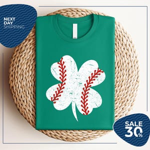 Könnte beinhalten: Smaragdgrünes T-Shirt mit einem weißen Baseball-Kleeblatt-Design und roten Nähten. Das Shirt wird auf einer gewebten Matte präsentiert. Der Text "NEXT DAY SHIPPING" ist oben links und "SALE 30%" unten rechts zu sehen.