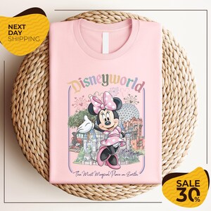 A pink t-shirt with a Minnie Mouse graphic and the text "Disneyworld" printed on it, placed on a woven basket.