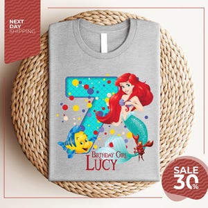 A gray t-shirt with a design featuring the character Ariel from the Disney movie "The Little Mermaid". The design includes the number 7 and the text "Birthday Girl Lucy".