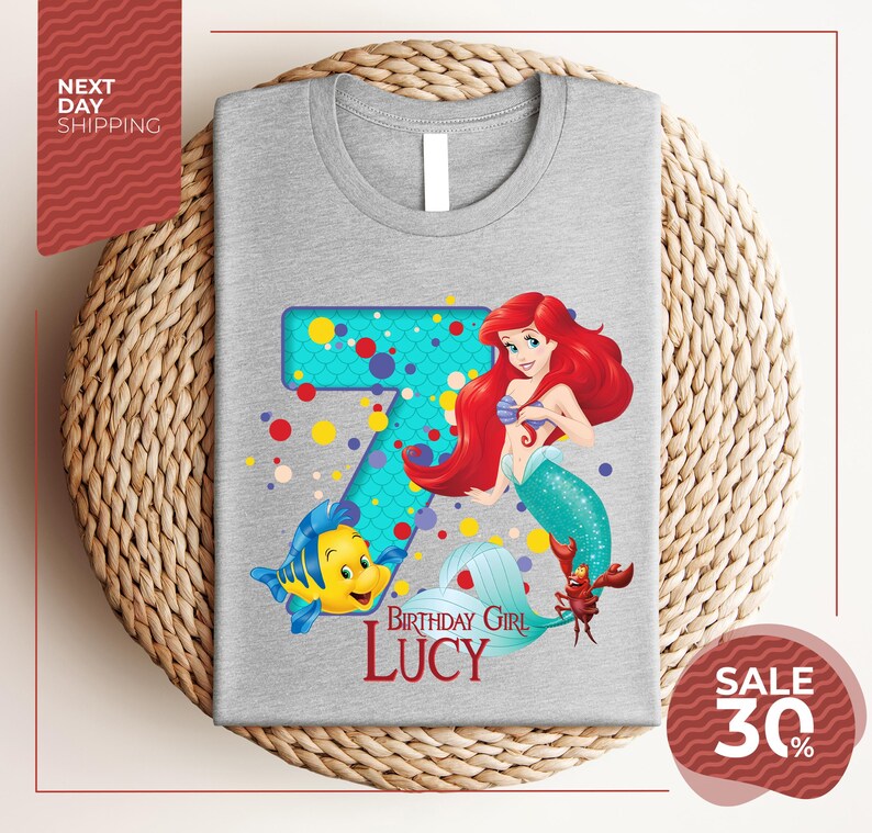 A gray t-shirt with a design featuring the character Ariel from the Disney movie "The Little Mermaid". The design includes the number 7 and the text "Birthday Girl Lucy".