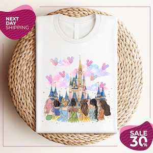Könnte beinhalten: Weißes T-Shirt mit Rundhalsausschnitt und einer farbenfrohen Illustration eines Märchenschlosses, Luftballons und einer Gruppe von Mädchen. Das Design umfasst rosa, blaue und gelbe Farbtöne. Das Shirt wird auf einer gewebten Oberfläche präsentiert.