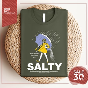 a green t-shirt with a graphic design depicting a person holding an umbrella and sprinkling salt on the ground. The text on the t-shirt reads "All these flavors and to be chosen are Salty", and there is a sale offer of "SALE 30%".