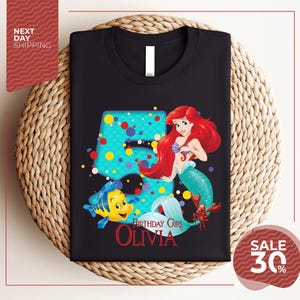 A black t-shirt with a design featuring the character Ariel from the Disney movie "The Little Mermaid". The design includes Ariel, a mermaid, and a fish, along with the text "Birthday Girl Olivia".