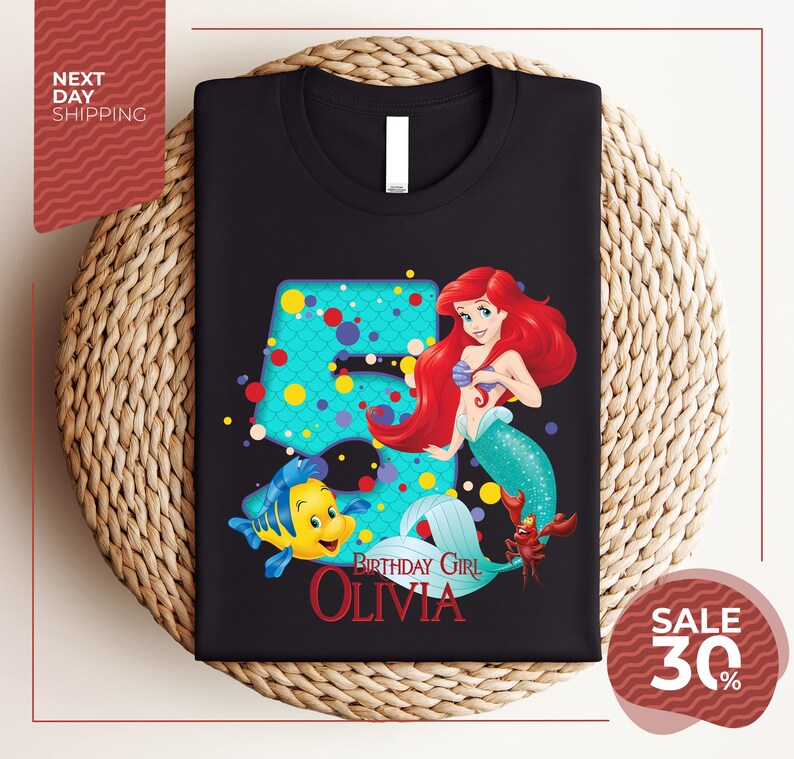 A black t-shirt with a design featuring the character Ariel from the Disney movie "The Little Mermaid". The design includes Ariel, a mermaid, and a fish, along with the text "Birthday Girl Olivia".