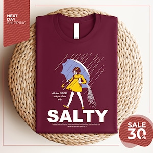 a maroon t-shirt with a graphic design depicting a woman holding an umbrella and sprinkling salt on the ground. The text on the t-shirt reads "ALL these flavors and to be chosen, SALTY" and "SALE 30%".