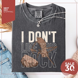 May include: Dark gray Comfort Colors t-shirt with the text "I DON'T LIKE THIS ROCK" and a cartoon insect holding a rock. A silver necklace is draped on the shirt. The shirt has a sale tag for 30% off.