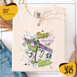 a white t-shirt with a colorful graphic of Buzz Lightyear from the Toy Story franchise, along with the text "Lightyear to Infinity and Beyond". The t-shirt is displayed on a white background, and there are some jeans and a necklace visible in the foreground.