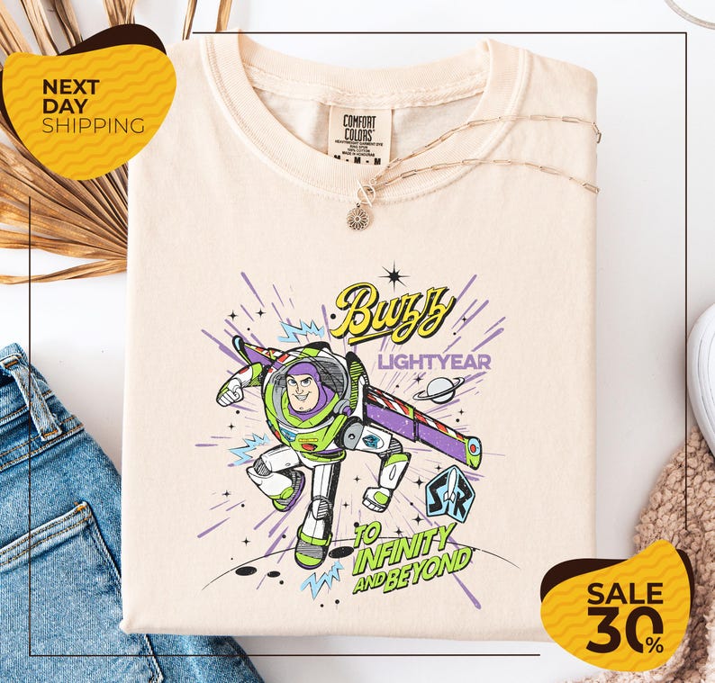 a white t-shirt with a colorful graphic of Buzz Lightyear from the Toy Story franchise, along with the text "Lightyear to Infinity and Beyond". The t-shirt is displayed on a white background, and there are some jeans and a necklace visible in the foreground.