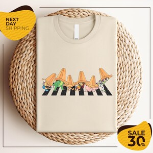 a beige t-shirt with a cartoon-style illustration of a crosswalk with cartoon characters wearing hats. The t-shirt is displayed on a woven basket, and there are two yellow sale signs in the image.