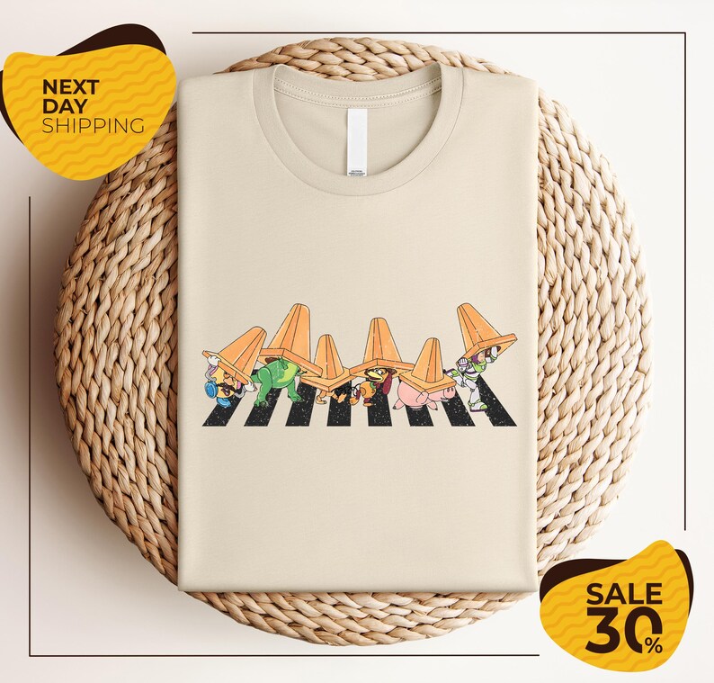 a beige t-shirt with a cartoon-style illustration of a crosswalk with cartoon characters wearing hats. The t-shirt is displayed on a woven basket, and there are two yellow sale signs in the image.