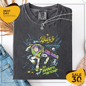 a gray t-shirt with a colorful graphic of Buzz Lightyear from the Toy Story franchise, along with the text "Buzz Lightyear to Infinity and Beyond". The t-shirt is displayed on a white background, and there are jeans and a necklace visible in the foreground.