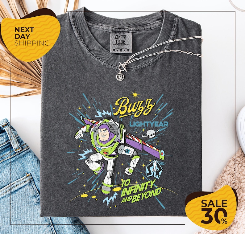 a gray t-shirt with a colorful graphic of Buzz Lightyear from the Toy Story franchise, along with the text "Buzz Lightyear to Infinity and Beyond". The t-shirt is displayed on a white background, and there are jeans and a necklace visible in the foreground.