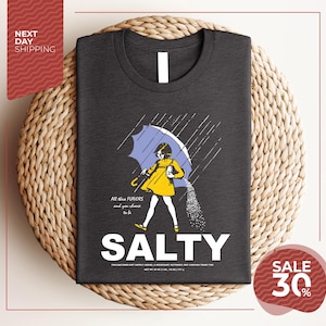 a gray t-shirt with a graphic design depicting a woman holding an umbrella and sprinkling salt on the ground. The text on the t-shirt reads "ALL these flavors and to be chosen, to be salty".