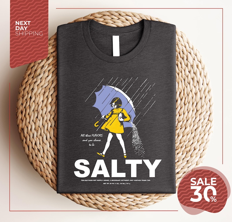a gray t-shirt with a graphic design depicting a woman holding an umbrella and sprinkling salt on the ground. The text on the t-shirt reads "ALL these flavors and to be chosen, to be salty".