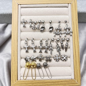 May include: A collection of silver-toned metal earrings and charms arranged on a beige velvet display tray. The set includes various cross designs, star shapes, small dangling charms, and four safety pin style earrings in gold and silver tones.