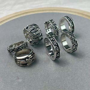 May include: A collection of six silver rings featuring intricate embossed designs, including floral scrollwork, cross motifs, and a dagger detail. The rings vary in width and thickness, with some displaying visible engravings on the inner bands.