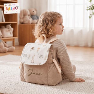 Puede incluir: Una mochila de pana beige con una solapa blanca que presenta un patrón estampado de pequeños caracoles, conejos y flores silvestres marrones. El frente de la bolsa está bordado con el nombre Jace en letra cursiva e incluye dos bolsillos laterales.