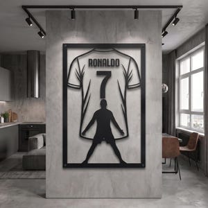 May include: A black metal wall art piece featuring a rectangular frame containing a silhouette of a soccer player standing in front of a jersey. The jersey displays the name RONALDO and the number 7 in bold, dark lettering centered on the back.