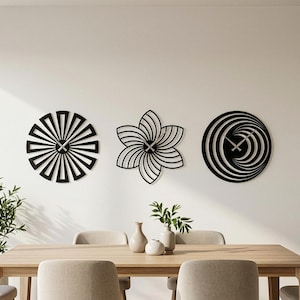 May include: Three black decorative wall clocks. One is a sunburst design, another is a flower, and the third is a spiral. The clocks are mounted on a white wall above a wooden dining table.