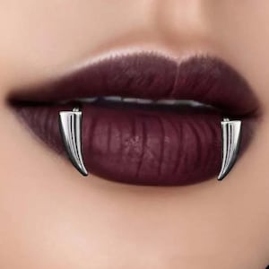 May include: A close-up shot of a person's lips, painted with dark burgundy lipstick. Two silver-coloured, fang-shaped lip piercings are attached to the lower lip, giving a vampire-like look. The image focuses on the mouth and the piercings.