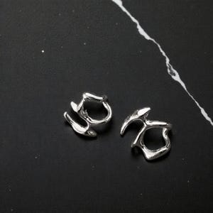 K&ouml;nnte beinhalten: Zwei silberfarbene Ohrringe in abstrakter Form. Jeder Ohrring hat ein einzigartiges, flie&szlig;endes Design mit abgerundeten und spitzen Elementen. Die Ohrringe werden auf einer dunklen, strukturierten Oberfl&auml;che pr&auml;sentiert, die mit ihrem gl&auml;nzenden Finish kontrastiert.