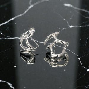 K&ouml;nnte beinhalten: Ein Paar silberfarbene Ohrringe in abstrakter Form. Die Ohrringe haben ein einzigartiges, skulpturales Design mit geschwungenen Linien und offenen R&auml;umen. Sie werden auf einer reflektierenden schwarzen Oberfl&auml;che mit wei&szlig;en Adern pr&auml;sentiert.