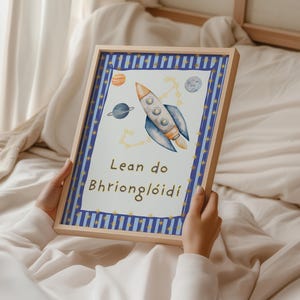 May include: A rectangular wooden frame containing a print with a blue and white striped border featuring gold stars. The center displays a watercolor rocket ship, planets, and constellations with the text Lean do Bhrionglóidí in brown lettering.