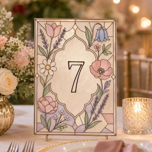 May include: A rectangular table number sign featuring a stained glass style design with a central number 7. The border is decorated with illustrations of pink tulips, blue bellflowers, daisies, and lavender sprigs on a cream-colored background.