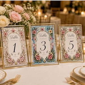Pode incluir: Três placas douradas com números de mesa com desenhos florais. Cada placa apresenta um número diferente: 1, 3 e 5. As placas têm um efeito de vitral com flores rosa, azuis e amarelas. A palavra "Table" está no topo de cada placa.