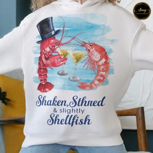 May include: White hoodie with a humorous design of two red lobsters clinking martini glasses. One lobster wears a top hat. The text "Shaken, Stirred & slightly Shellfish" is printed below the image.