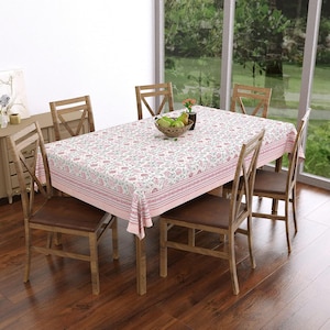 Pode incluir: Uma mesa de jantar retangular com uma toalha de mesa branca com um padrão floral rosa e verde e uma borda listrada. A mesa está rodeada por quatro cadeiras de madeira com assentos castanhos. Uma taça com peras verdes está no centro.