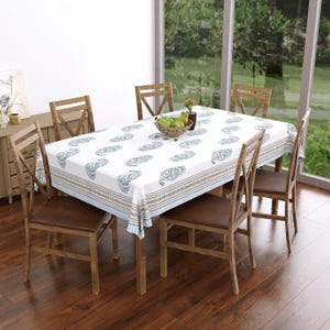 Peut inclure: Une table à manger rectangulaire recouverte d'une nappe blanche avec un motif cachemire bleu répétitif et une bordure rayée. La table est entourée de quatre chaises en bois à assise marron, avec un bol de poires vertes au centre.
