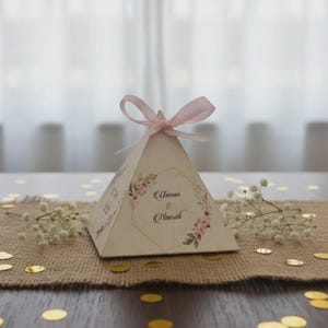 May include: A pyramid-shaped favor box with a pink ribbon bow. The box is decorated with floral designs and the names "Usman & Himrah". The box sits on a burlap surface with gold confetti and small white flowers.