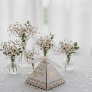May include: A pyramid-shaped gift box with a white ribbon sits on a table, surrounded by small vases of baby's breath. The box has gold accents and text. The overall theme is elegant and celebratory.