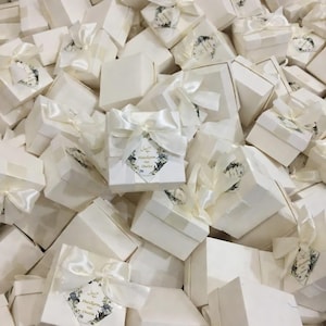 Puede incluir: Una colección de cajas de regalo blancas, cada una con un lazo de cinta de raso. Cada caja cuadrada tiene una etiqueta con detalles florales y texto. Las cajas están dispuestas en una pila, sugiriendo una gran cantidad de regalos.