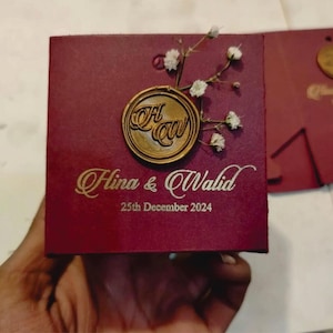Puede incluir: Caja de invitación de boda burdeos con detalles dorados. Presenta un sello de cera dorado con iniciales y delicadas flores blancas. Los nombres "Hina & Walid" y la fecha "25 de diciembre de 2024" están impresos en dorado.
