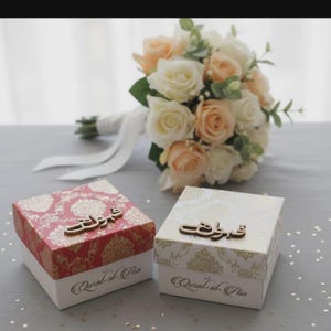 Puede incluir: Dos cajas de regalo decorativas con patrones florales y escritura árabe, una roja y dorada, la otra crema y dorada. Un ramo de rosas blancas y melocotón con vegetación se encuentra en el fondo. Las cajas tienen el texto "Qural-ul-Atin".
