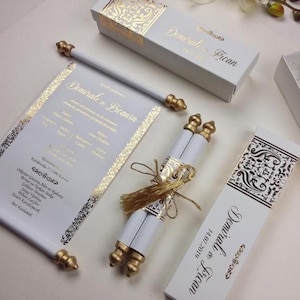 Puede incluir: Juego de invitaciones de boda blanco con detalles dorados. Incluye un pergamino, cajas y una tarjeta, todos con escritura dorada y patrones decorativos. El conjunto está diseñado para un evento especial.