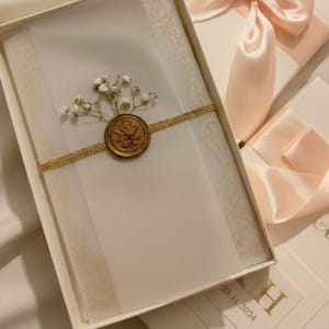 May include: A white invitation card with a gold seal and a sprig of white flowers, presented in a white box. The card is secured with a gold ribbon. The box is decorated with a light pink ribbon.
