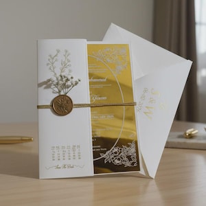 Może przedstawiać: Elegancki zestaw zaproszeń ślubnych z białą kartą ze złotymi akcentami, złotą wkładką i pasującą kopertą. Zaproszenie zdobi kwiatowy wzór, pieczęć woskowa i kalendarz. Na kopercie widnieją inicjały "M & W" oraz data ślubu.