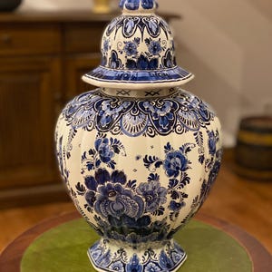 Könnte beinhalten: Eine große, weiße Keramikvase mit blauem Blumendesign und passendem Deckel. Die Vase hat einen bauchigen Körper und einen ausgestellten Sockel. Der Deckel ist mit einer kleinen, blauen Spitze versehen. Die Vase ist mit aufwendigen blauen Blumenmustern verziert.