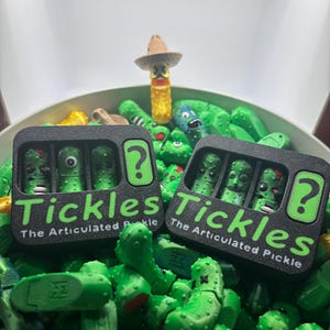Könnte beinhalten: Eine Ansammlung grüner, beweglicher Gurkenspielzeuge, einige mit Gesichtern, und zwei schwarze Schachteln mit der Aufschrift "Tickles The Articulated Pickle". Eine Gurke trägt einen Sombrero. Die Schachteln enthalten jeweils drei Gurkenfiguren.